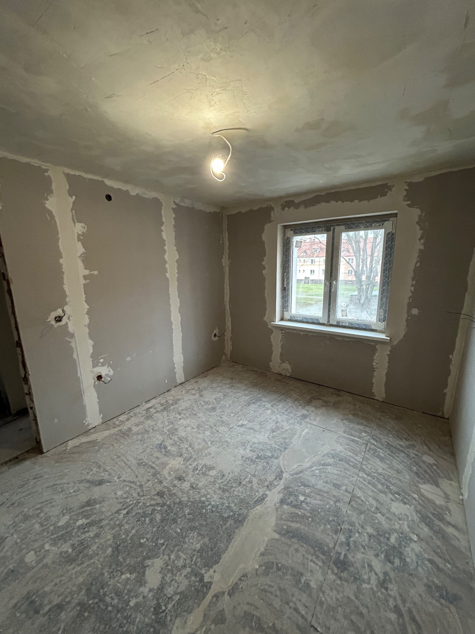 Pokój w trakcie szpachlowania ścian i sufitu, widoczne gładzie gipsowe, okno z widokiem na budynki. W tle żarówka na kablu. Podłoga zabezpieczona folią.