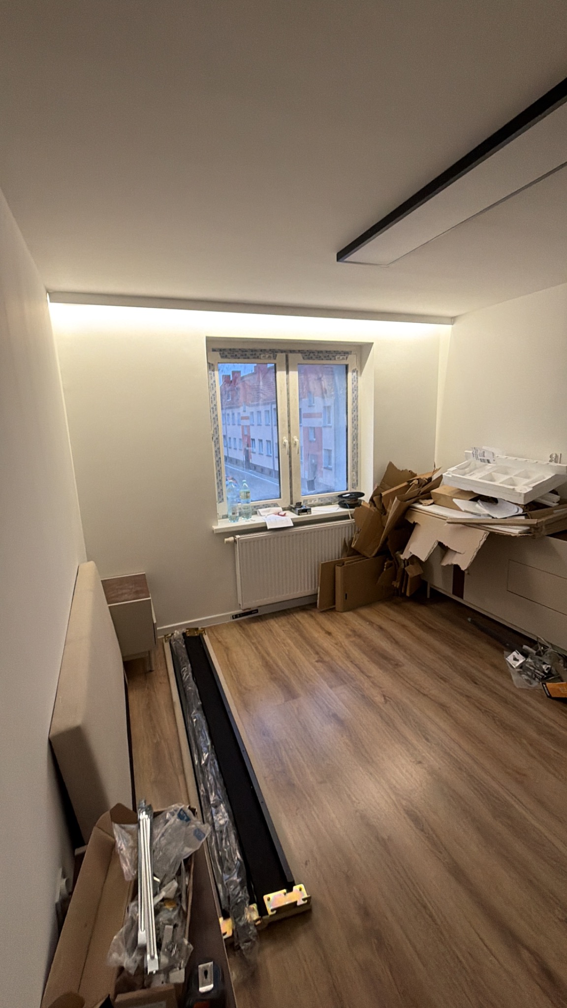 Pokój w trakcie przygotowań do remontu: panele podłogowe, okno z widokiem na budynki, kartony i elementy wyposażenia. Oświetlenie sufitowe.