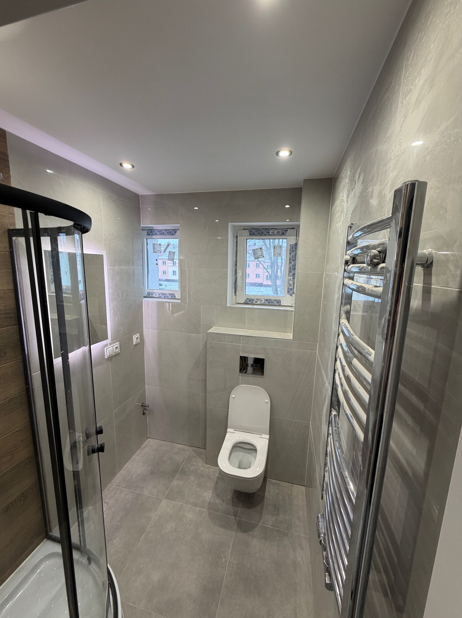 Jasna łazienka z szarymi płytkami, kabiną prysznicową z czarnymi ramami, toaletą i grzejnikiem drabinkowym. Dwa okna z widokiem na zimowy krajobraz.