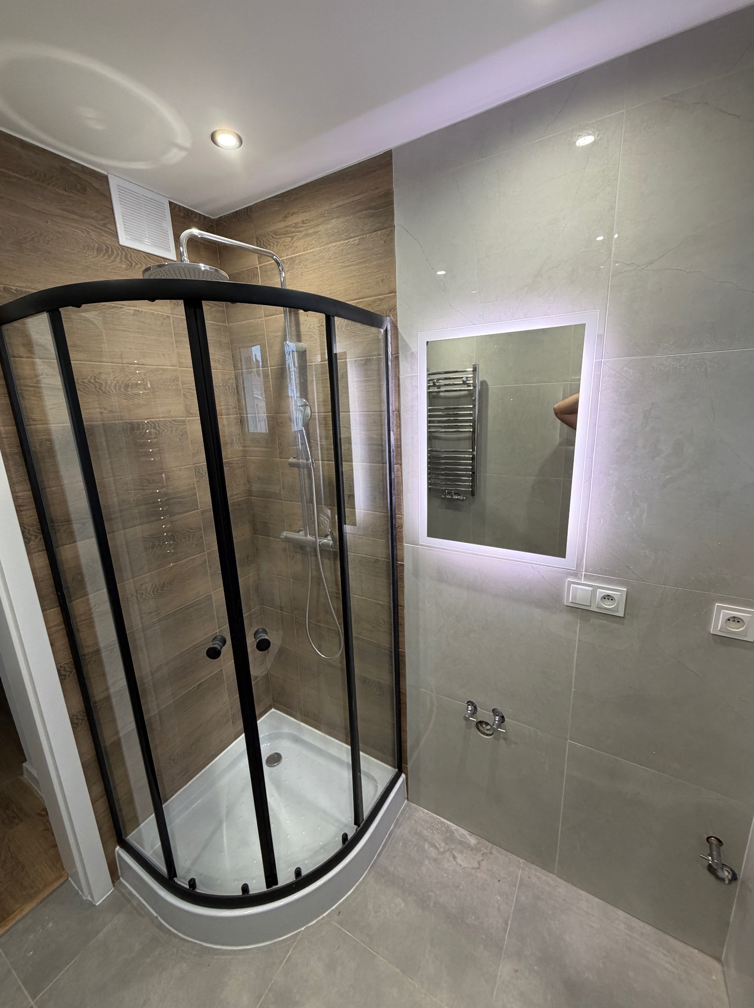 Nowoczesna łazienka z kabiną prysznicową w czarnej ramie, drewnopodobnymi płytkami i podświetlanym lustrem. Widoczne szare płytki i minimalistyczne wykończenie.