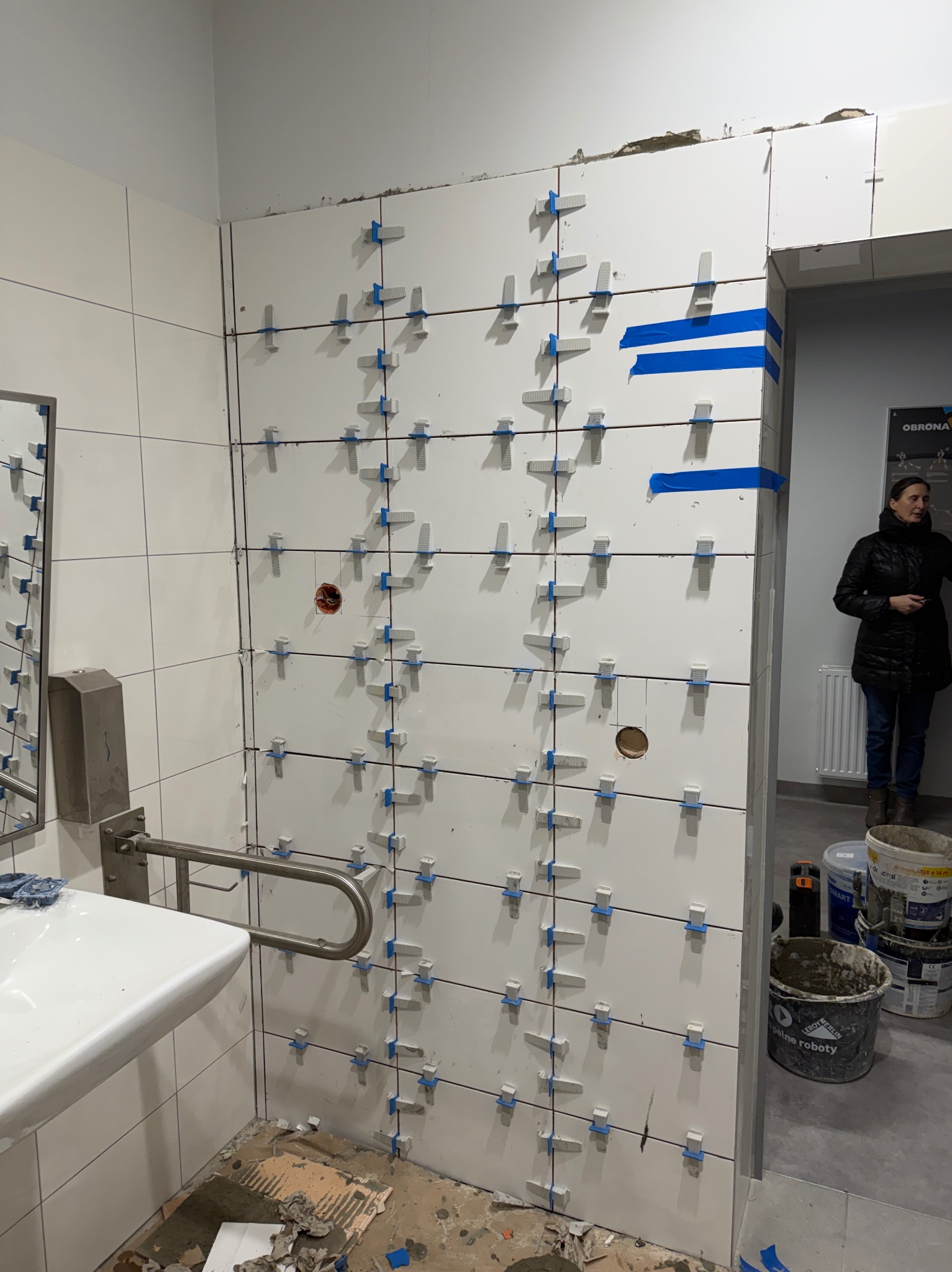 Białe płytki z niebieskimi krzyżykami dystansowymi na ścianie łazienki w trakcie montażu. Widoczne narzędzia i osoba w tle. Etap prac glazurniczych.