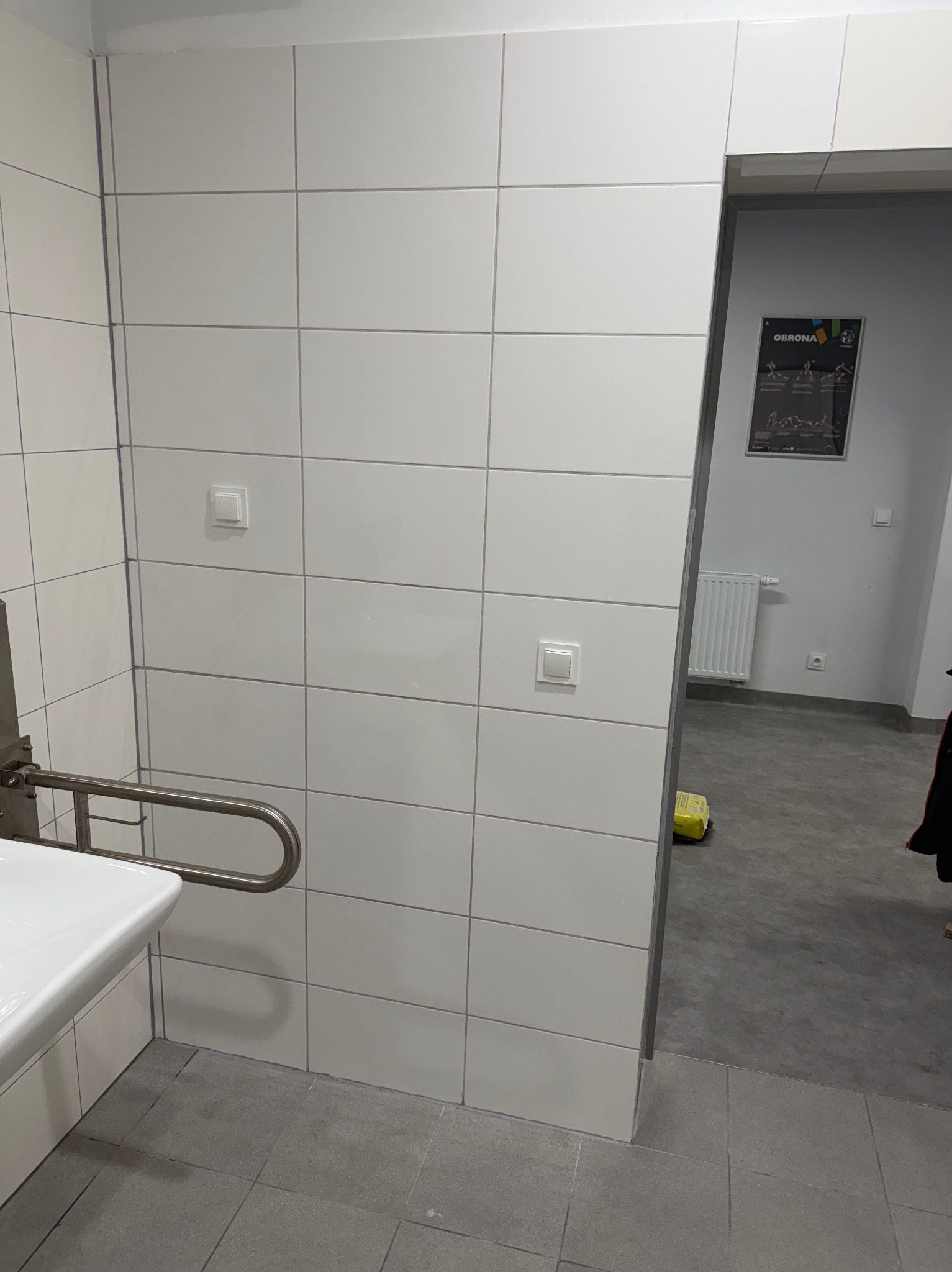 Wykończona łazienka z białymi płytkami, uchwytem dla niepełnosprawnych i widocznym przejściem do innego pomieszczenia. Minimalistyczny design, neutralna kolorystyka.