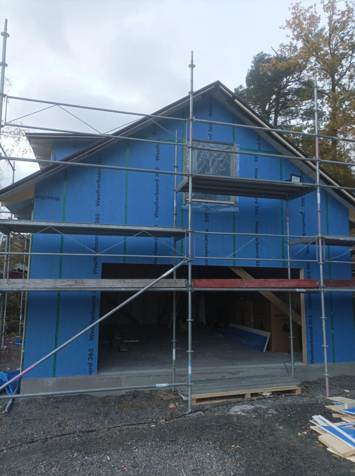 Budowa domu w trakcie: niebieska membrana na ścianach, rusztowania, widoczny garaż. Etap przed wykończeniem elewacji, jesienna sceneria w tle.
