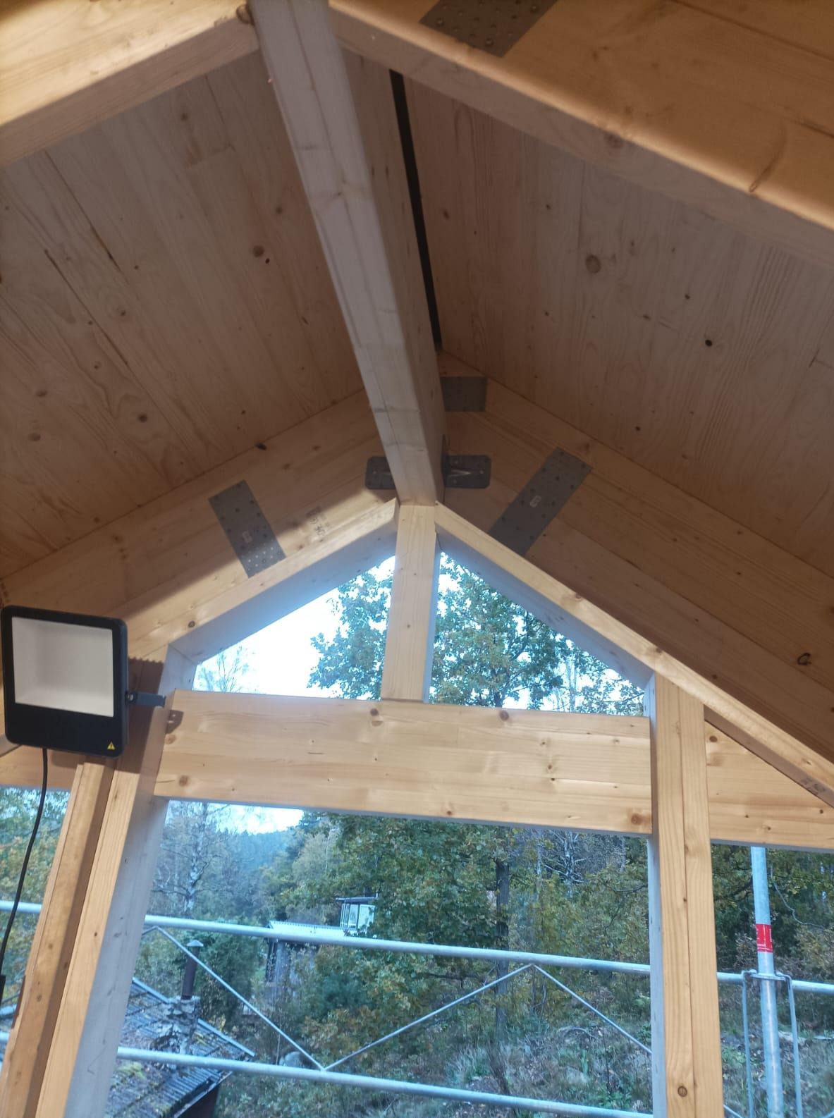 Wnętrze konstrukcji domu w trakcie budowy, widoczne drewniane belki i metalowe łączniki, fragment okna z widokiem na drzewa i reflektor budowlany.