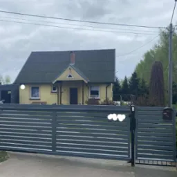 Nowoczesna, pozioma brama wjazdowa w kolorze grafitowym, z przesuwnym skrzydłem, na tle domu jednorodzinnego i zieleni. Widoczna skrzynka na listy i tabliczka 'Ja tu pilnuję'.