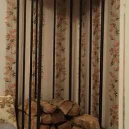 Metalowy stojak na drewno z drewnem w środku, na górze dekoracje, obok kanapa z poduszką, w tle tapeta w liście.