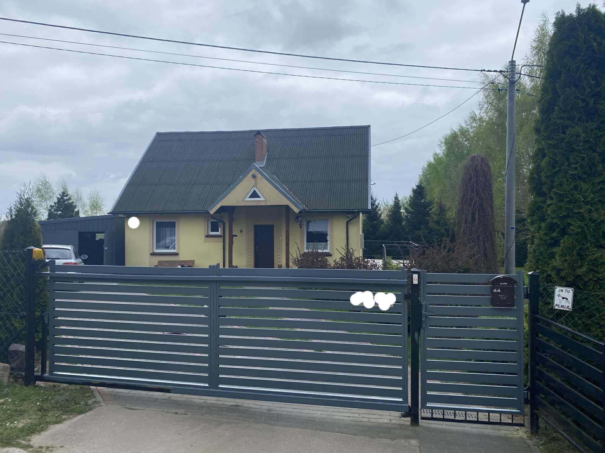 Nowoczesna, pozioma brama wjazdowa w kolorze grafitowym, z przesuwnym skrzydłem, na tle domu jednorodzinnego i zieleni. Widoczna skrzynka na listy i tabliczka 'Ja tu pilnuję'.