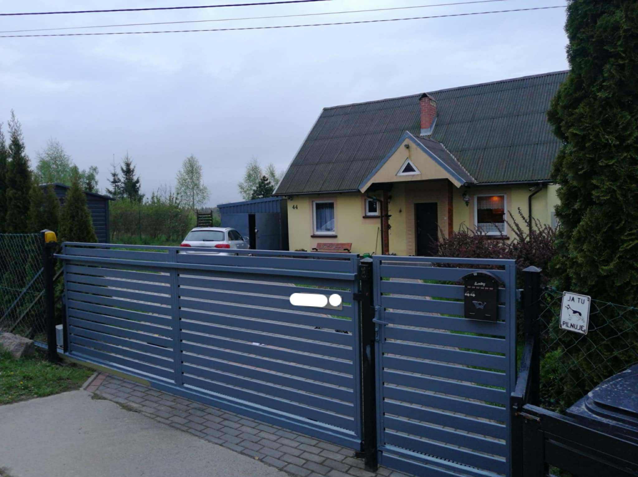 Nowoczesna, pozioma brama wjazdowa w kolorze grafitowym, z widocznym domem w tle. Na bramie skrzynka pocztowa i tabliczka z psem.