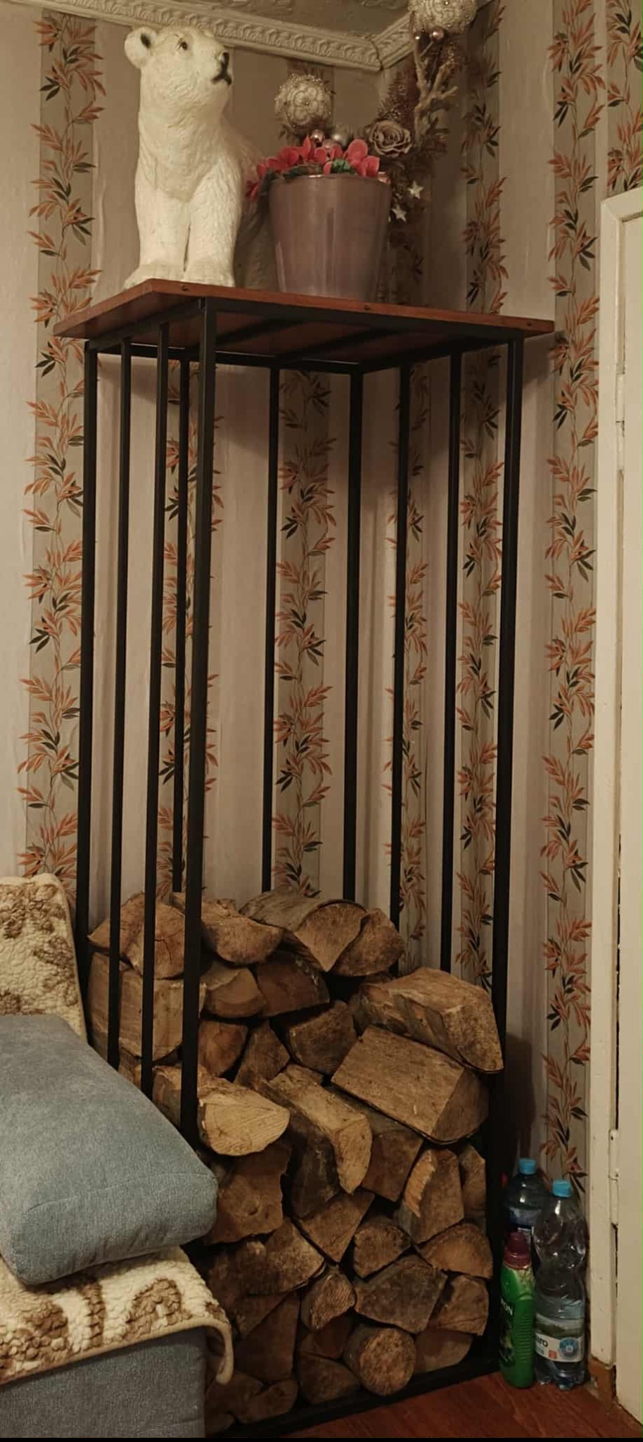 Metalowy stojak na drewno z drewnem w środku, na górze dekoracje, obok kanapa z poduszką, w tle tapeta w liście.