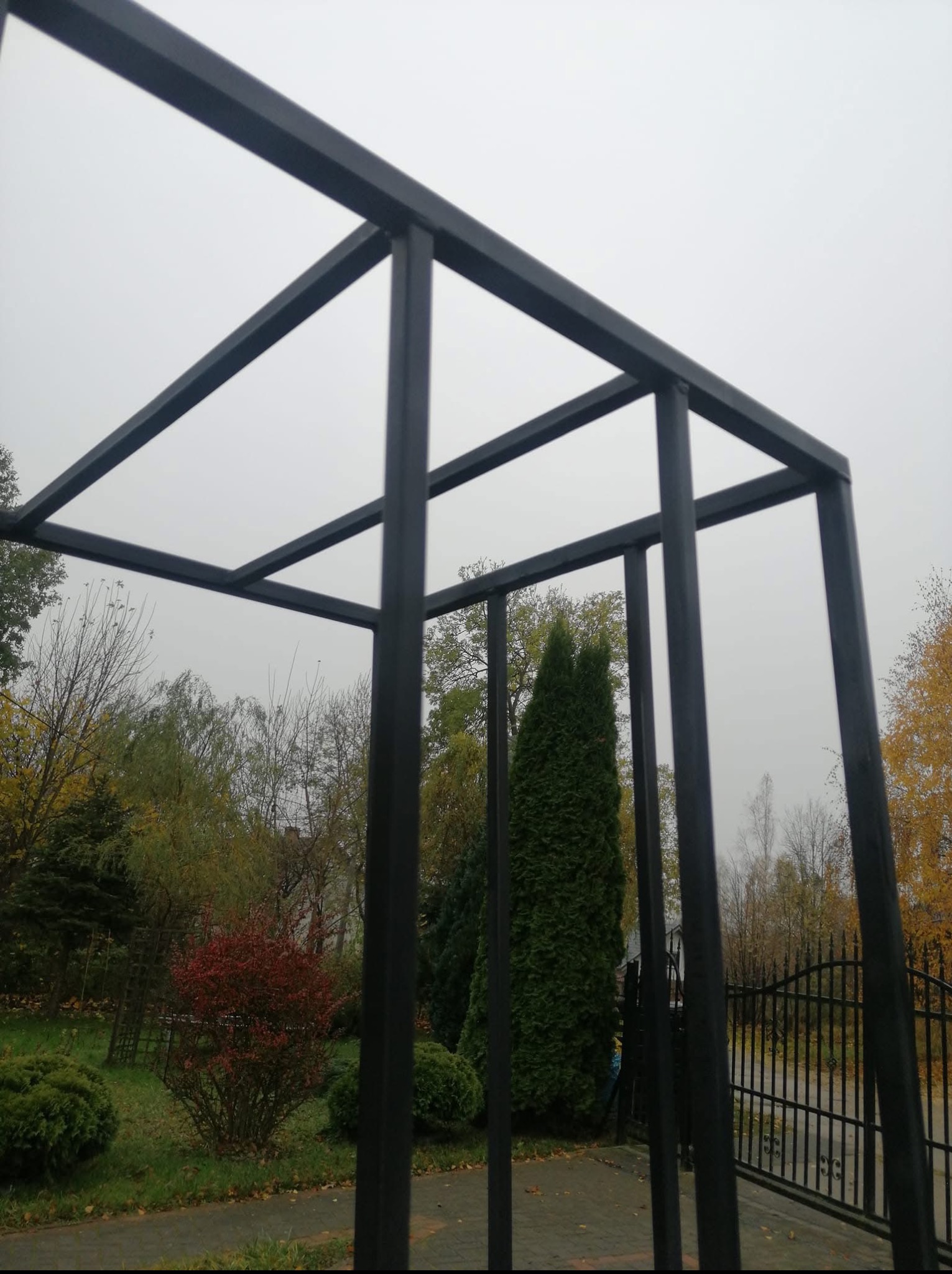 Metalowa konstrukcja w Warczu, prawdopodobnie pergola lub element dekoracyjny ogrodu. Widok na zieleń i niebo. Geometryczny design, czarny kolor.