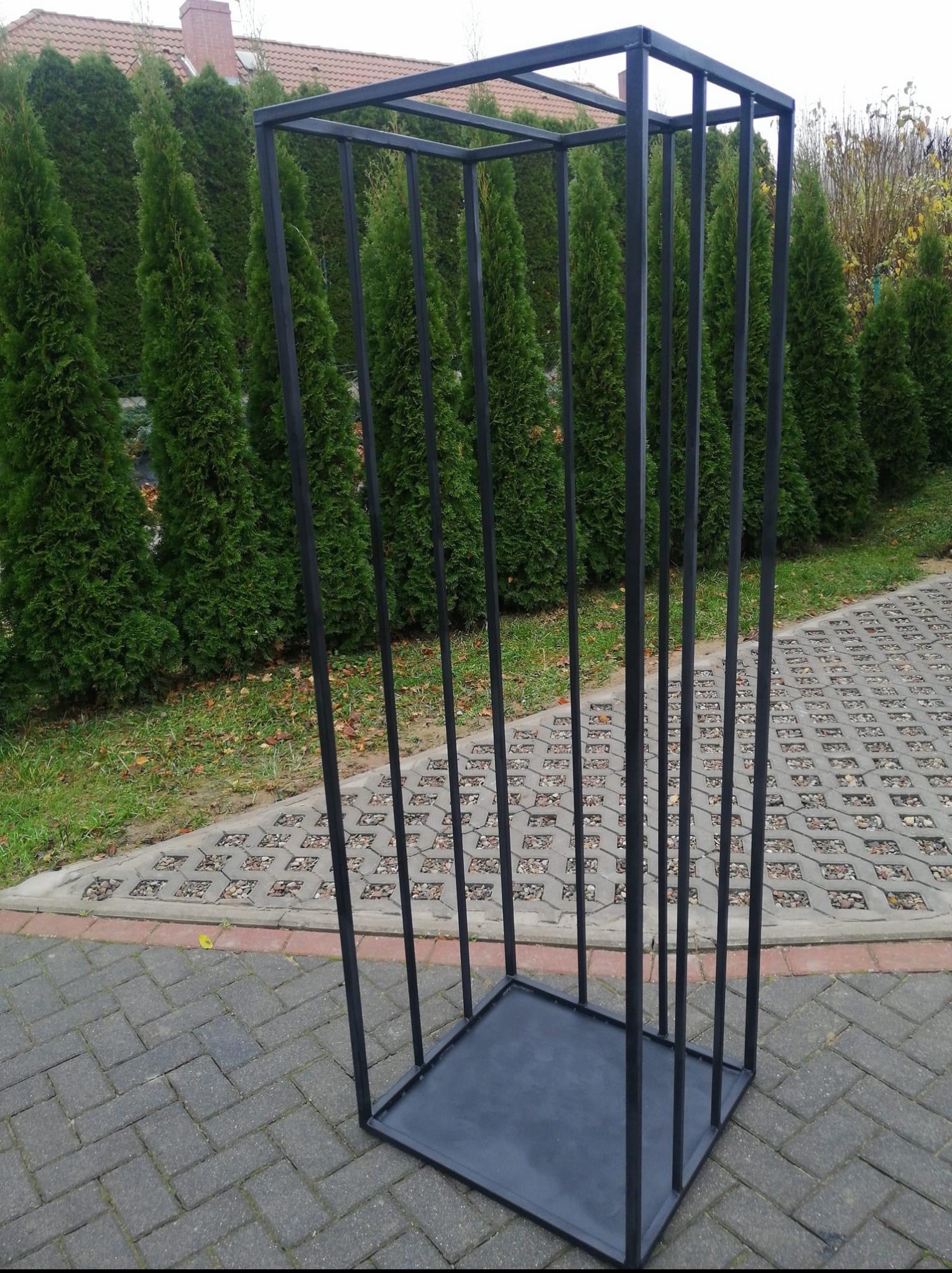 Czarna, metalowa konstrukcja prostopadłościenna z pionowymi prętami, umieszczona na kostce brukowej, z tłem zielonych tuj. Nietypowy element dekoracyjny.