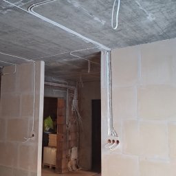 Sparky - Surowy stan: białe kable elektryczne prowadzone po suficie i ścianach w nowym budynku, widoczne puszki instalacyjne.  Przewody pod podłogą.