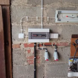 Szara, surowa ściana z ceglanymi wstawkami. Zamontowana biała skrzynka elektryczna, przełącznik i gniazda. Widoczne kable w białych listwach. Całość sprawia wrażenie instalacji w garażu.