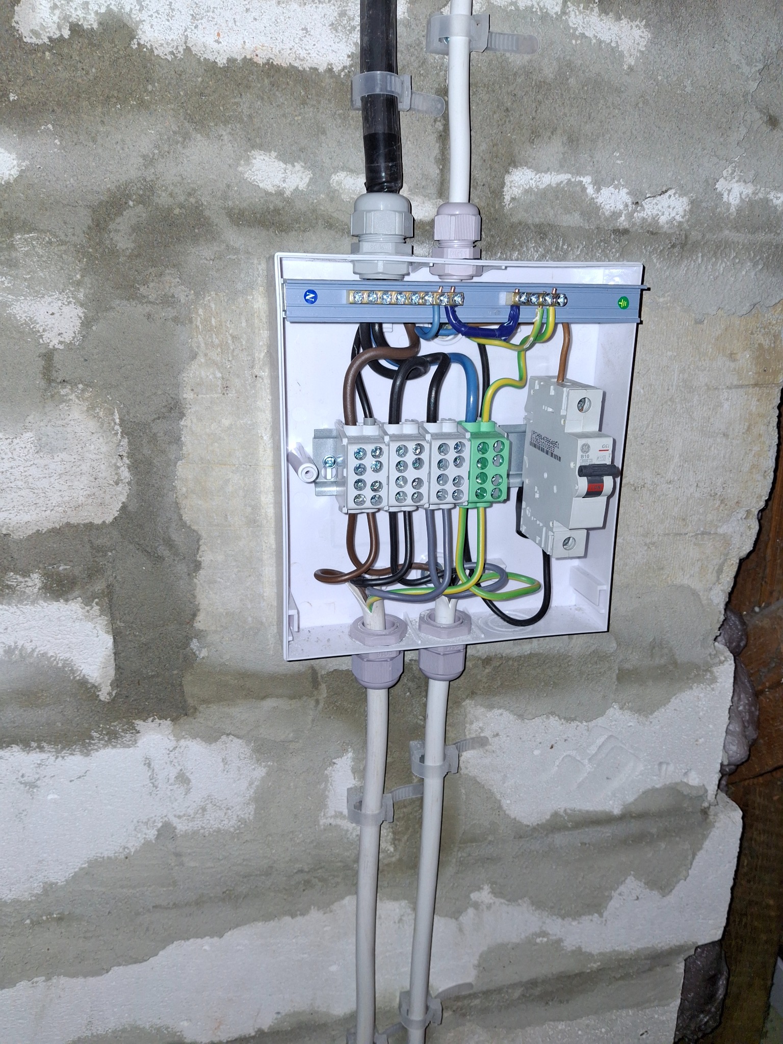 Otwarta skrzynka elektryczna z podłączonymi przewodami i zabezpieczeniami, zamontowana na surowej ścianie z betonu komórkowego. Widoczne kable w peszlach.