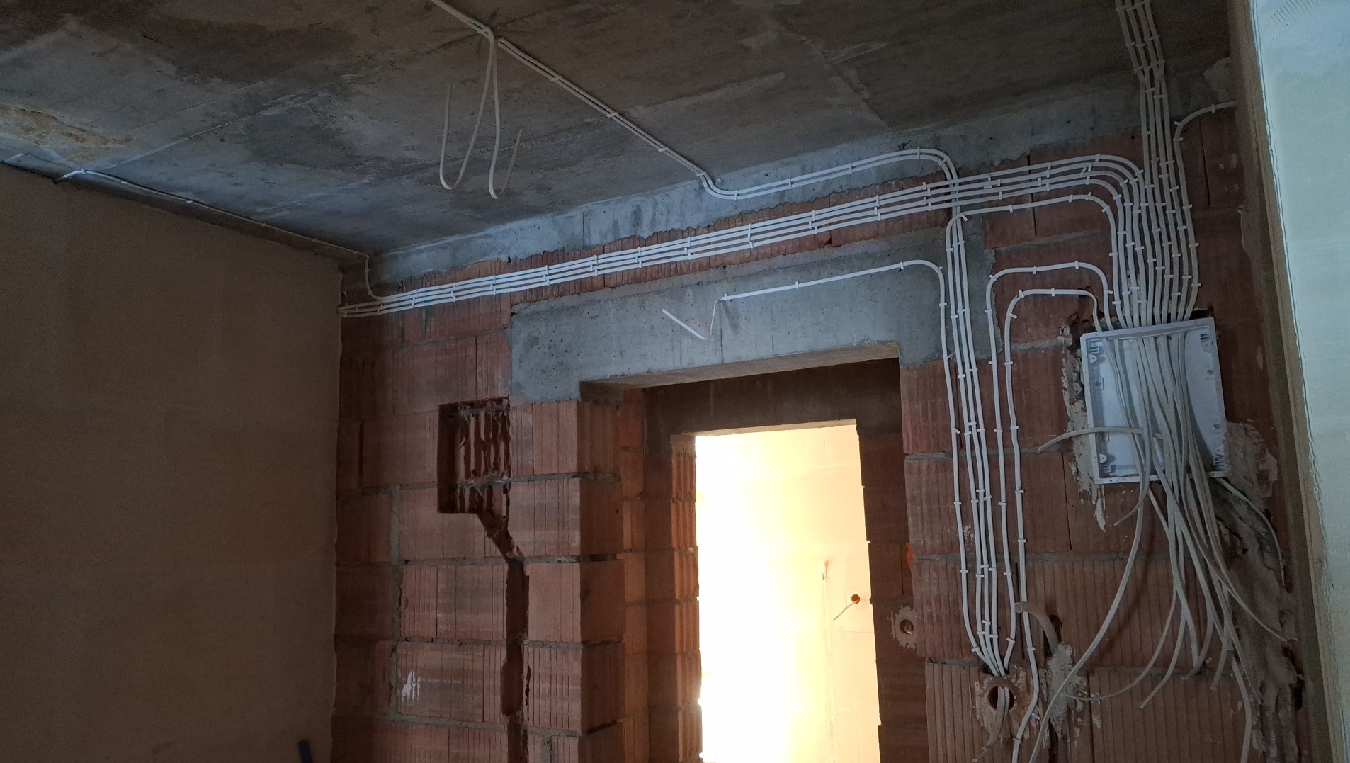 Surowy stan: białe kable elektryczne prowadzone po ceglanej ścianie i betonowym suficie w nowym budynku, skrzynka elektryczna z wiszącymi kablami.