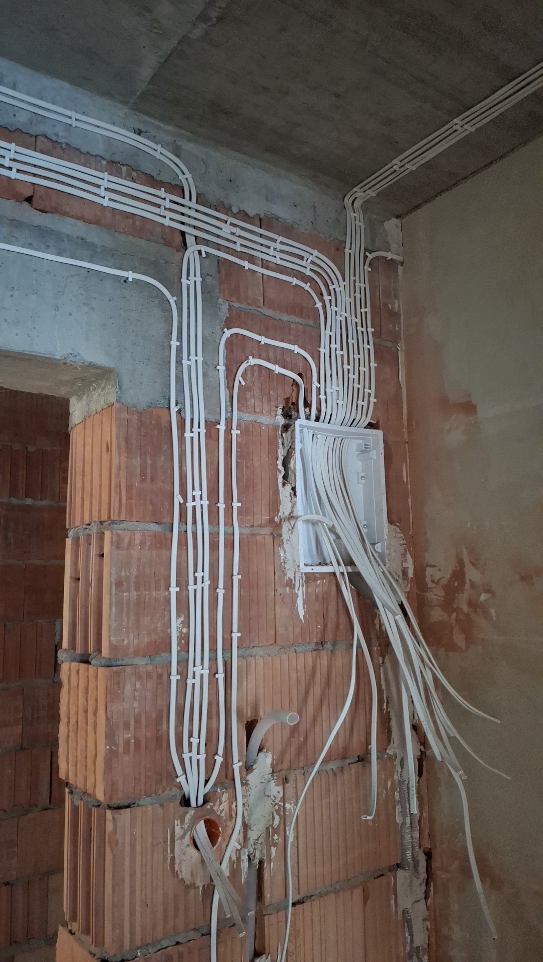 Białe rurki instalacji elektrycznej, prowadzone po ceglanej ścianie w surowym wnętrzu. Widoczna skrzynka rozdzielcza z wystającymi przewodami. Ściany w trakcie wykończenia.