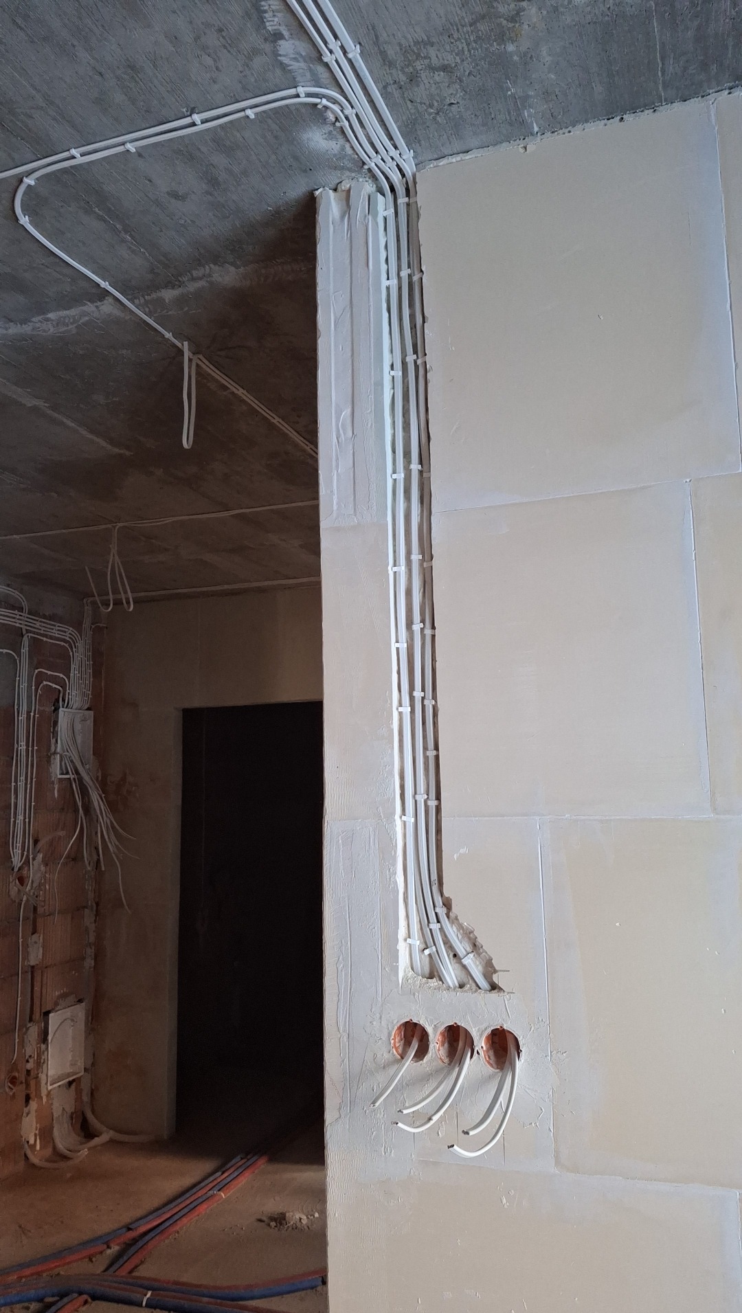Białe kable elektryczne w peszlach, prowadzone po ścianie i suficie w surowym wnętrzu. Widoczne puszki instalacyjne i przewody. Etap prac instalacyjnych.