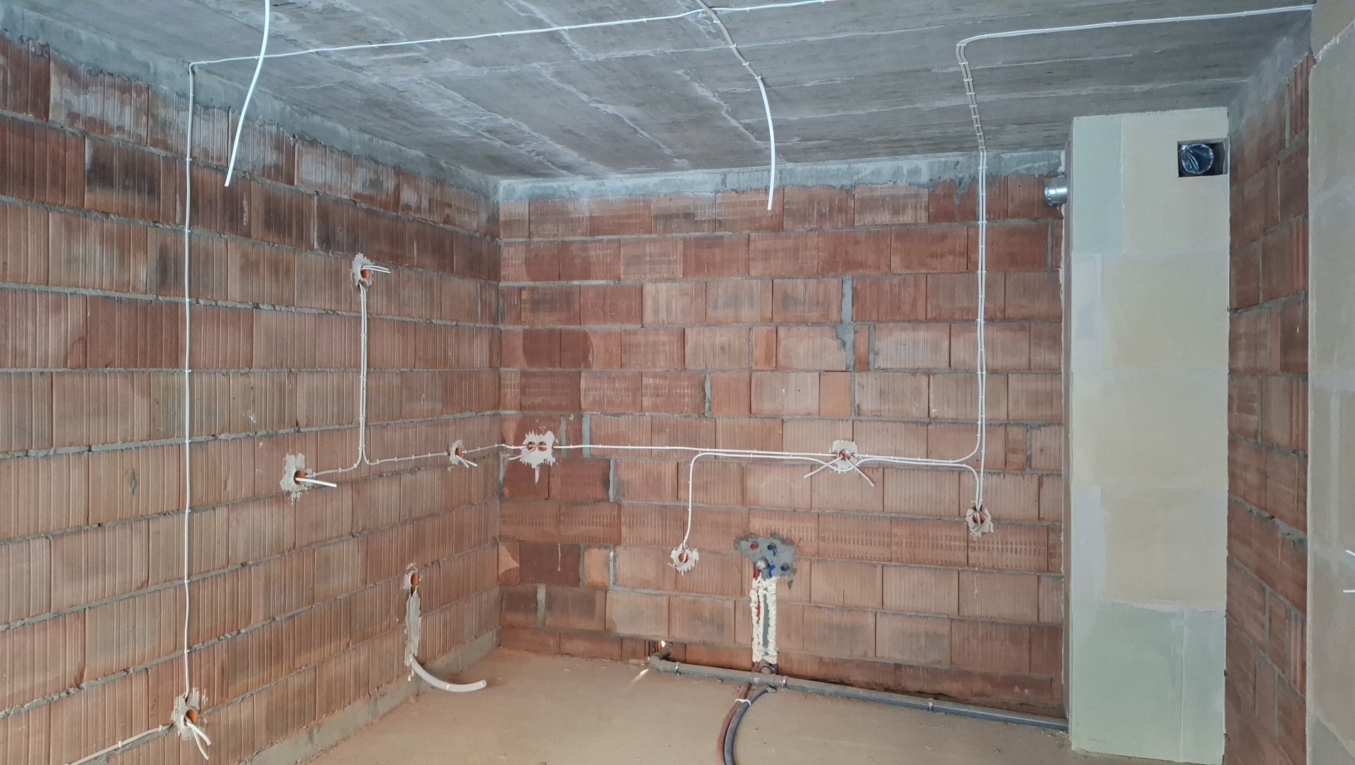 Wnętrze w trakcie instalacji elektrycznej: widoczne kable prowadzone po ceglanej ścianie i pod sufitem, puszki instalacyjne, fragment wentylacji. Surowy stan wykończenia.