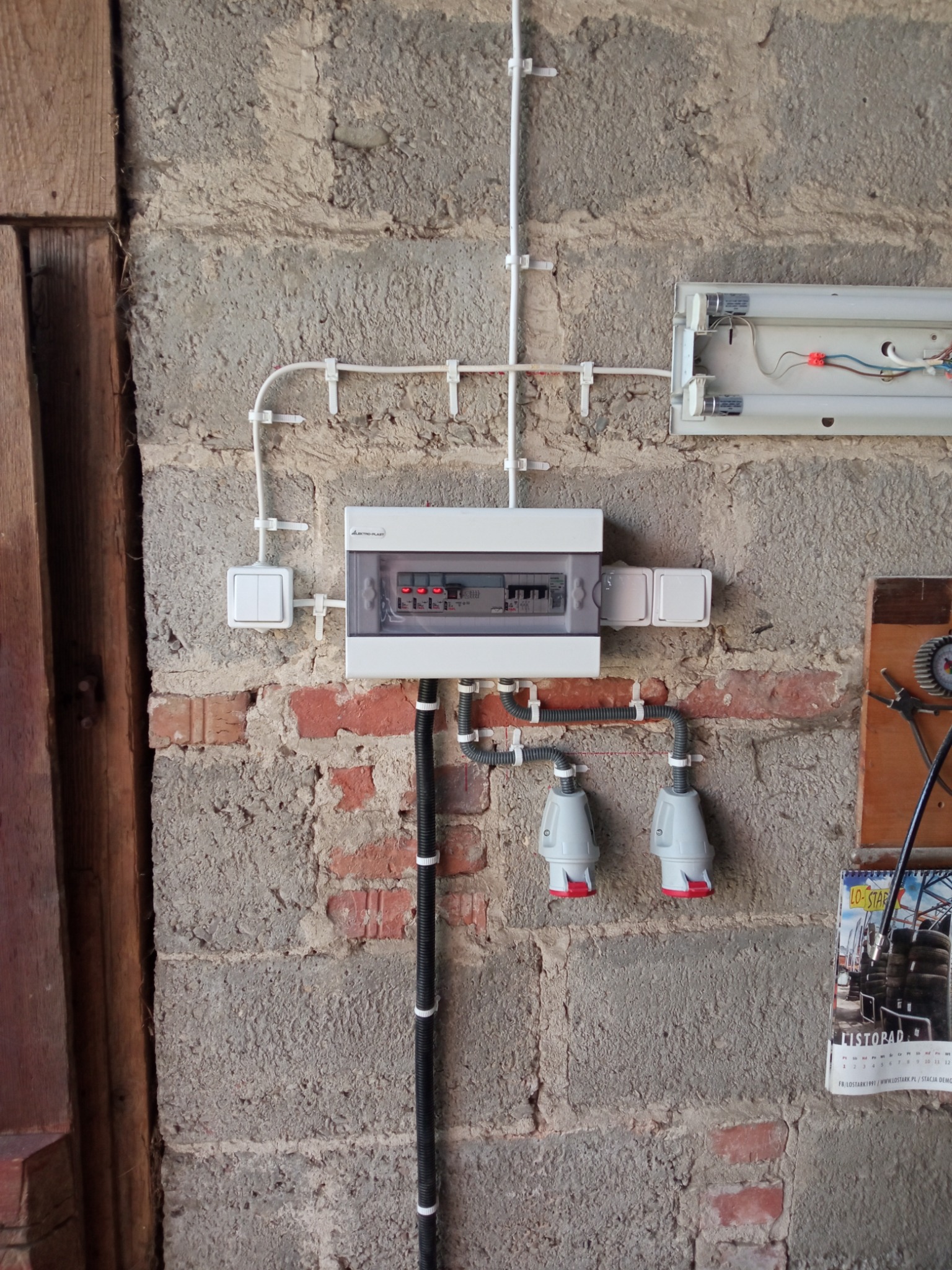 Szara, surowa ściana z ceglanymi wstawkami. Zamontowana biała skrzynka elektryczna, przełącznik i gniazda. Widoczne kable w białych listwach. Całość sprawia wrażenie instalacji w garażu.