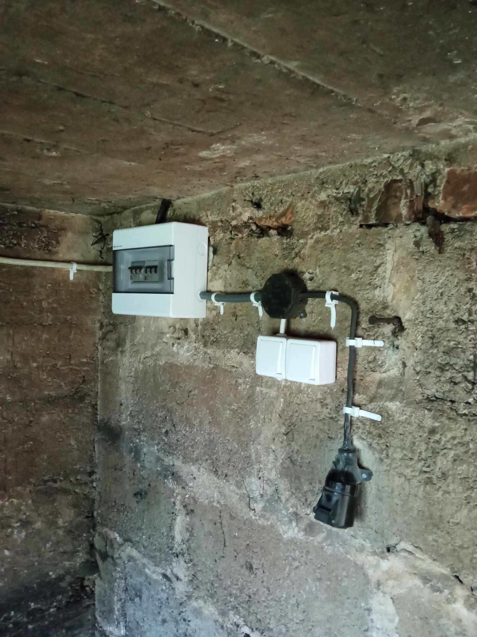 Instalacja elektryczna: biała skrzynka rozdzielcza, puszka instalacyjna i podwójny włącznik na surowej, ceglano-kamiennej ścianie. Widoczne kable i uchwyty mocujące.