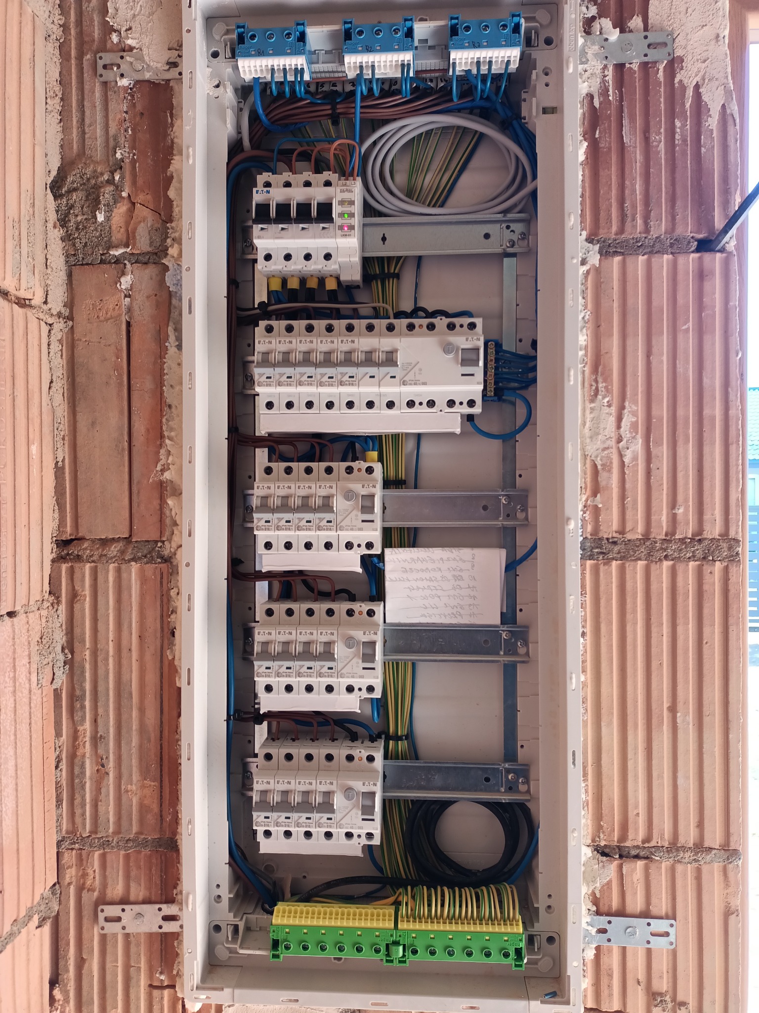 Rozdzielnica elektryczna z zainstalowanymi bezpiecznikami i przewodami, zamontowana w surowej ścianie z cegły dziurawki. Widoczne elementy mocujące i notatka z parametrami.