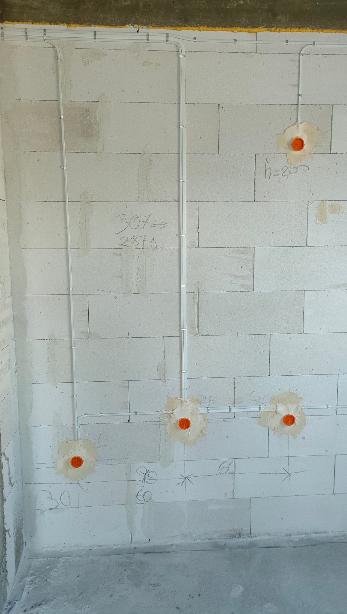 Montaż przewodów elektrycznych w białej ścianie z betonu komórkowego, widoczne pomarańczowe puszki instalacyjne i oznaczenia wysokości na ścianie.