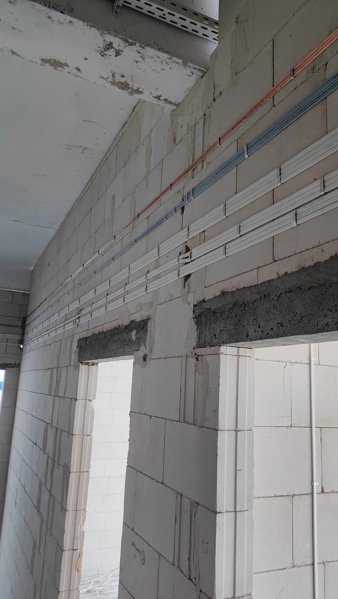 Ściana z betonu komórkowego z poziomymi rurami instalacji elektrycznej w kolorach miedzi, szarym i białym, zamocowanymi do ściany. Widoczne otwory drzwiowe.