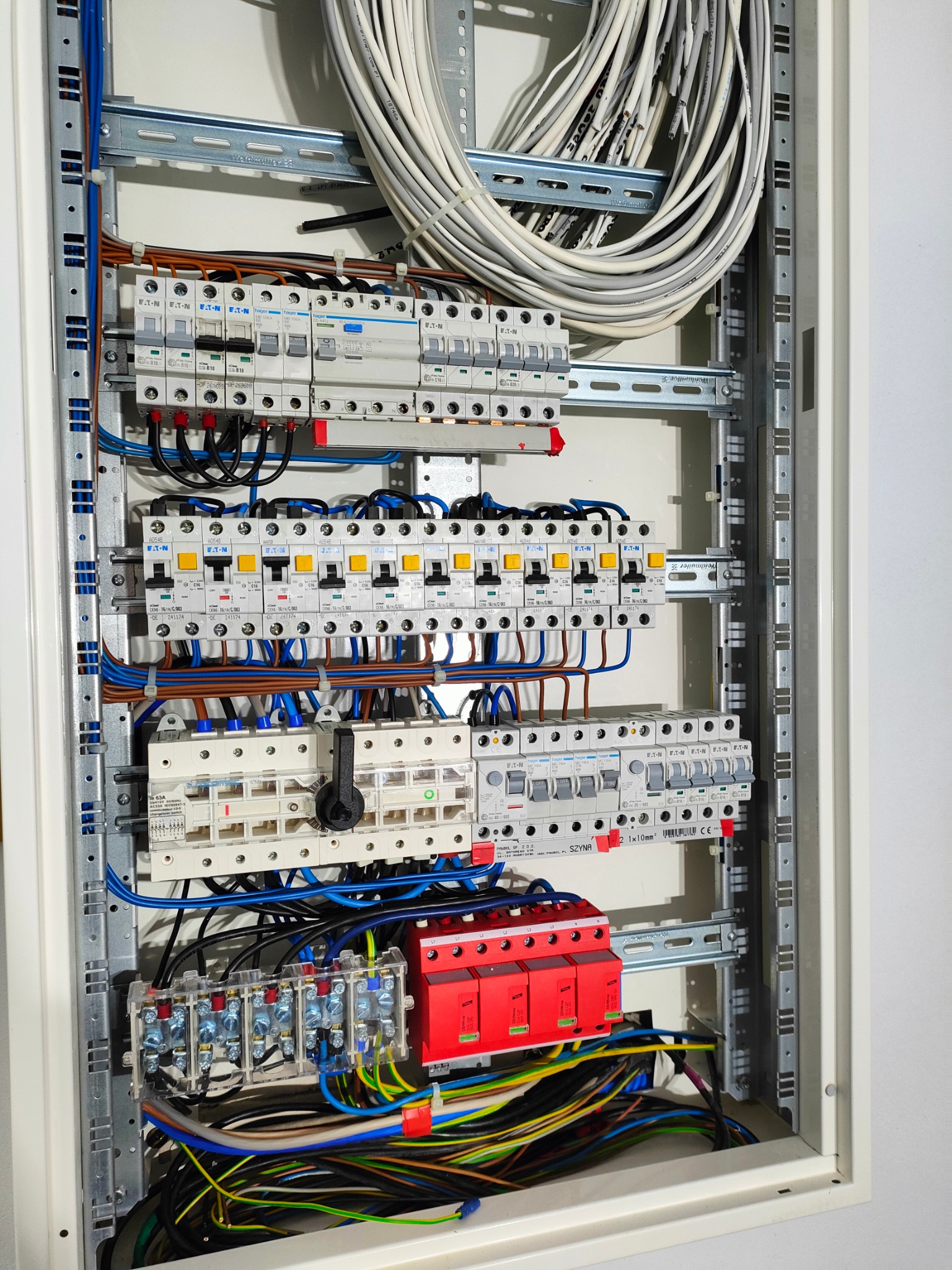Rozdzielnica elektryczna z zamontowanymi bezpiecznikami, przewodami w różnych kolorach i szyną. Widoczne elementy montażowe i oznaczenia na urządzeniach.