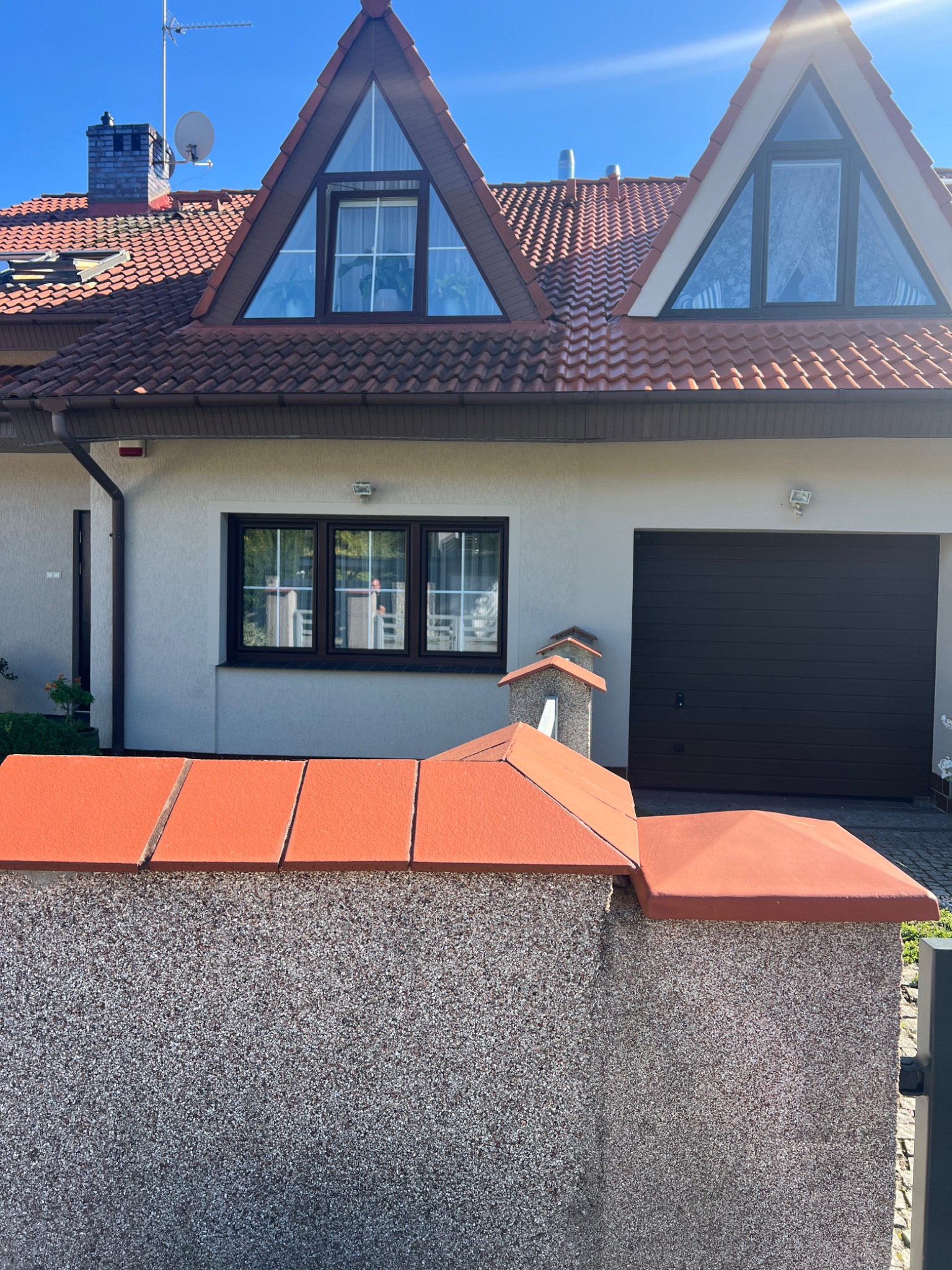 Dom jednorodzinny z czerwonym dachem i elewacją w jasnym kolorze. Widoczny fragment ogrodzenia z elementami w kolorze dachu. Słoneczny dzień.