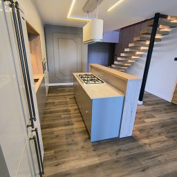 Markos usługi stolarskie Marcin Kołos - Nowoczesna kuchnia z wyspą i drewnianymi schodami. Oświetlenie LED podkreśla minimalistyczny design. Fronty szafek w odcieniach szarości, drewniana podłoga.