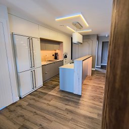 Markos usługi stolarskie Marcin Kołos - Nowoczesna kuchnia z wyspą, fronty w odcieniach szarości, drewniany blat i podłoga, oświetlenie LED. Widok z perspektywy wejścia, fragment zasłony na pierwszym planie.