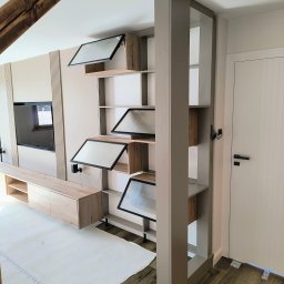 Markos usługi stolarskie Marcin Kołos - Nowoczesna szafa z półkami i szklanymi drzwiczkami, wkomponowana w aranżację salonu z drewnianą belką stropową i panelem TV. Widoczne zabezpieczenia podłogi.