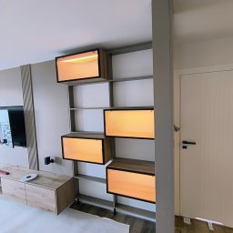 Markos usługi stolarskie Marcin Kołos - Nowoczesna szafa wnękowa z podświetlanymi półkami i elementami drewnopodobnymi, zamontowana w jasnym wnętrzu. Widoczny fragment mebla RTV i drzwi.