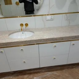 Biała szafka łazienkowa z blatem z jasnego kamienia, złotymi detalami i kryształowymi gałkami, odbicie osoby w lustrze. Nowoczesny design.