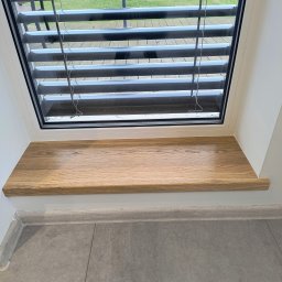 Markos usługi stolarskie Marcin Kołos - Jasny, dębowy parapet wewnętrzny z zaokrąglonym brzegiem, zamontowany pod oknem z żaluzjami poziomymi. Szara podłoga z płytek i białe ściany.