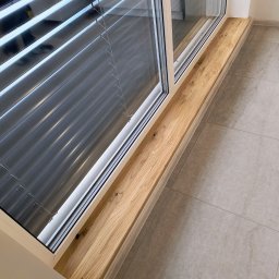 Markos usługi stolarskie Marcin Kołos - Jasny, drewniany parapet z wyraźnym usłojeniem, wkomponowany w nowoczesne okno z żaluzjami, na tle szarej, kwadratowej podłogi z płytek. Detal wnętrza.