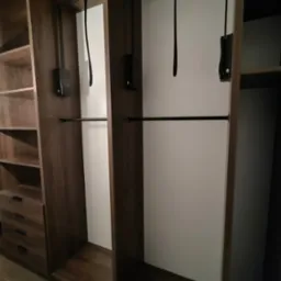 Wnętrze szafy wnękowej z ciemnego drewna, z drążkami na ubrania i półkami. Widoczne mechanizmy opuszczania drążków.