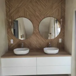 Nowoczesna łazienka z drewnianą mozaiką na ścianie, okrągłymi lustrami i białymi umywalkami nablatowymi. Wiszące lampy dodają elegancji.