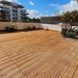 Słoneczny taras z desek kompozytowych, z widokiem na budynki mieszkalne. Drobne donice z roślinami i minimalistyczne meble ogrodowe dopełniają aranżację.