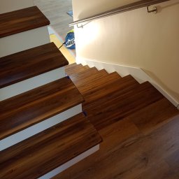 Markos usługi stolarskie Marcin Kołos - Drewniane stopnie schodów z metalową poręczą w Ełku. Widok z góry na dół, ciepłe oświetlenie podkreśla naturalny odcień drewna, białe podstopnice.