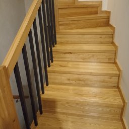 Markos usługi stolarskie Marcin Kołos - Jasne, dębowe schody z czarnymi, pionowymi balustradami, widok z dołu. Naturalne oświetlenie podkreśla strukturę drewna i kontrast z ciemnymi elementami.