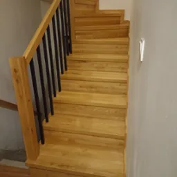 Jasne, dębowe schody z czarną, metalową balustradą w nowoczesnym domu. Widoczne detale wykończenia i naturalna tekstura drewna.