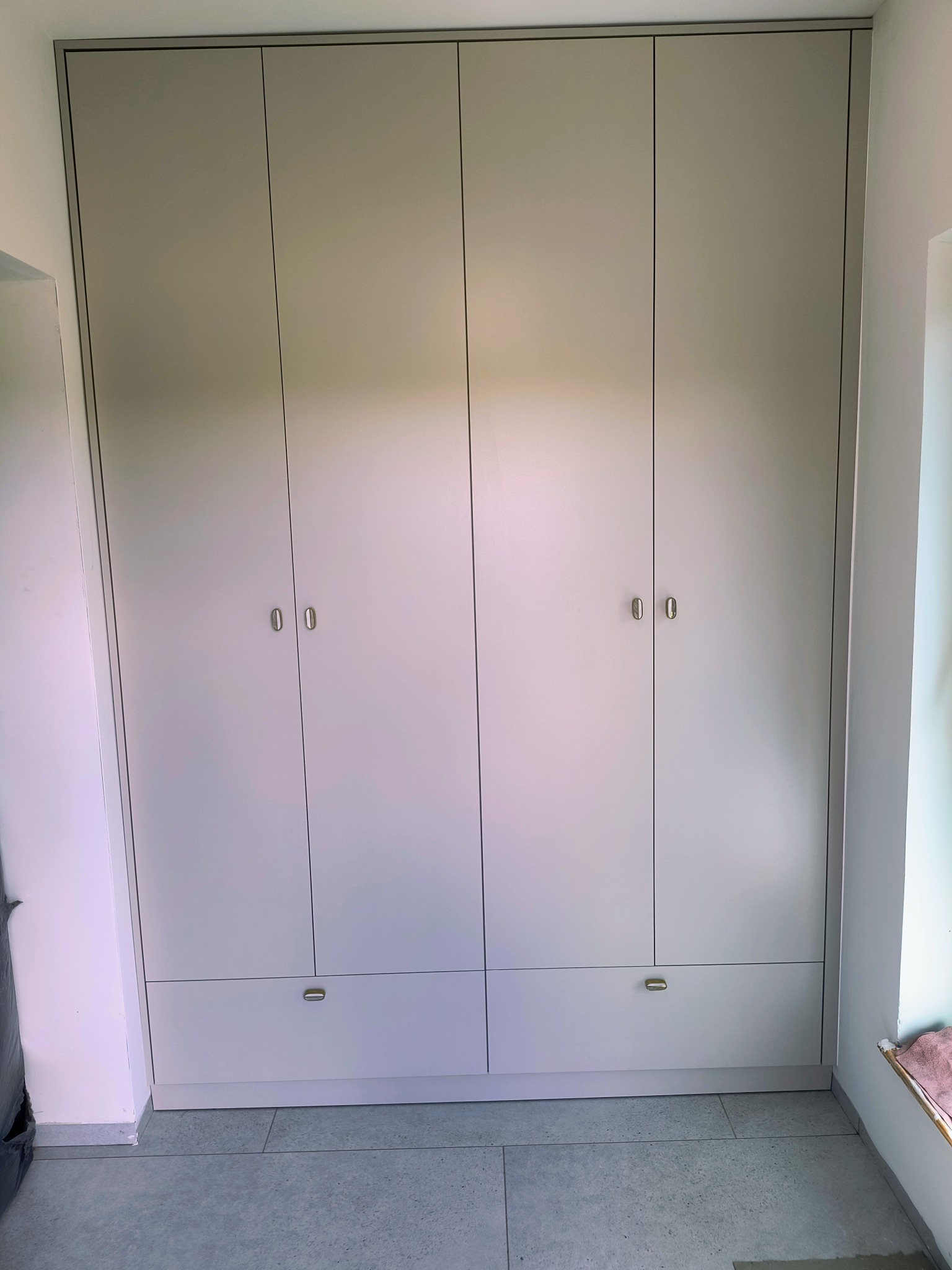 Czterodrzwiowa szafa wnękowa z szufladami w odcieniach szarości, z minimalistycznymi uchwytami, wkomponowana w jasne wnętrze. Widok z przodu, na podłodze szare płytki.