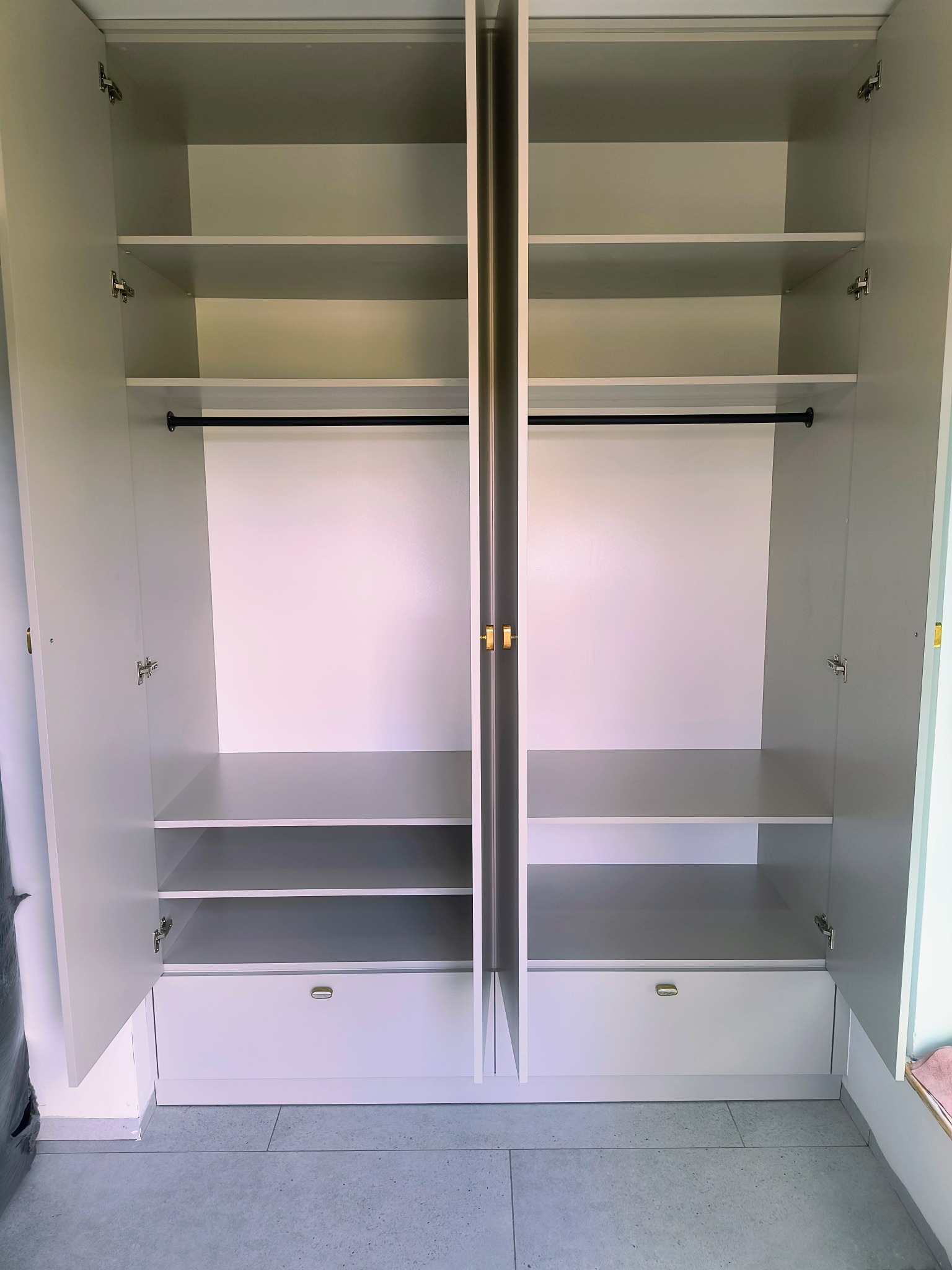 Wnętrze otwartej, jasnej szafy wnękowej z półkami, drążkiem na ubrania i szufladami. Widoczne zawiasy i minimalistyczne uchwyty. Podłoga wyłożona szarymi płytkami.