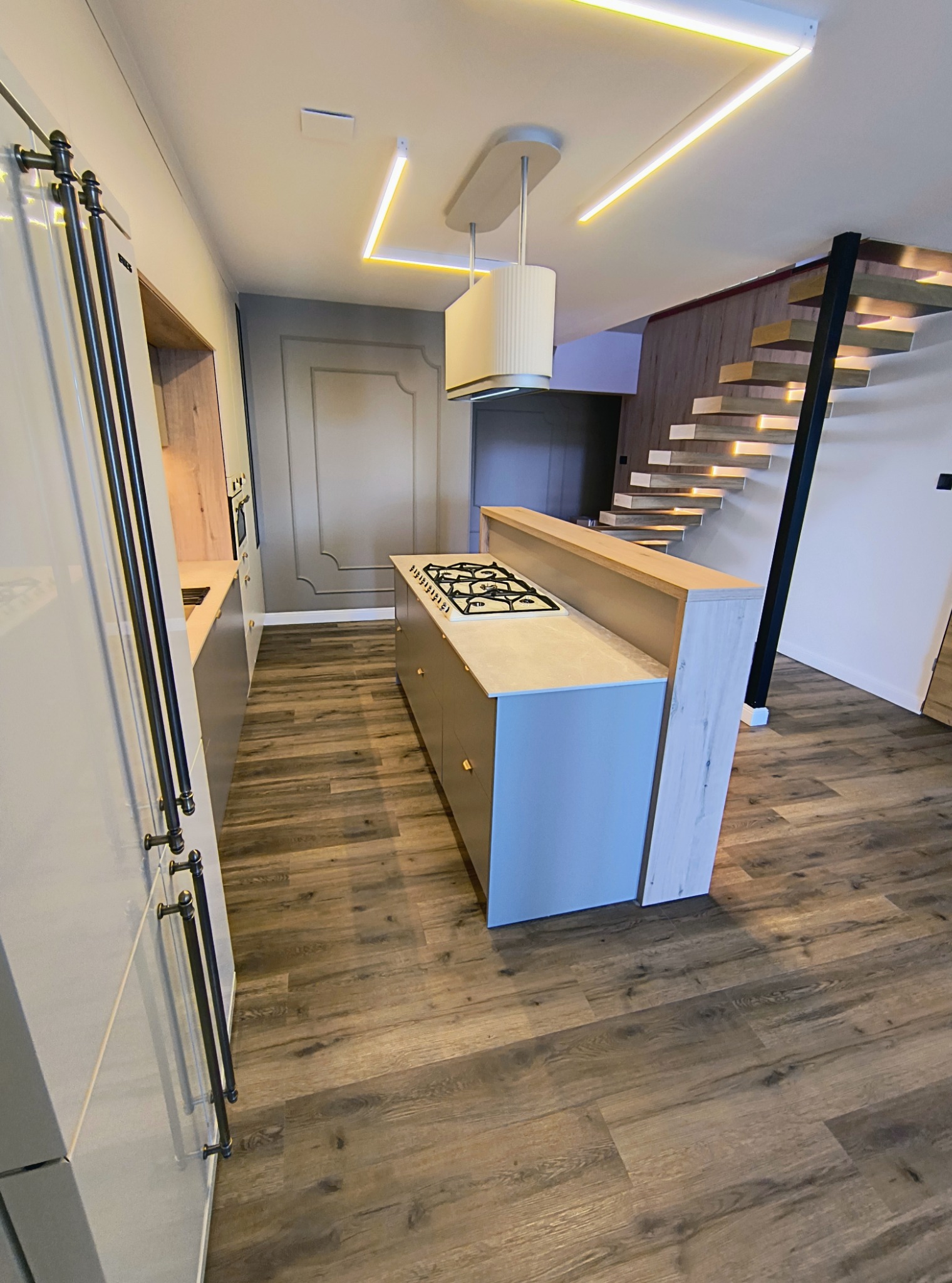 Nowoczesna kuchnia z wyspą i drewnianymi schodami. Oświetlenie LED podkreśla minimalistyczny design. Fronty szafek w odcieniach szarości, drewniana podłoga.