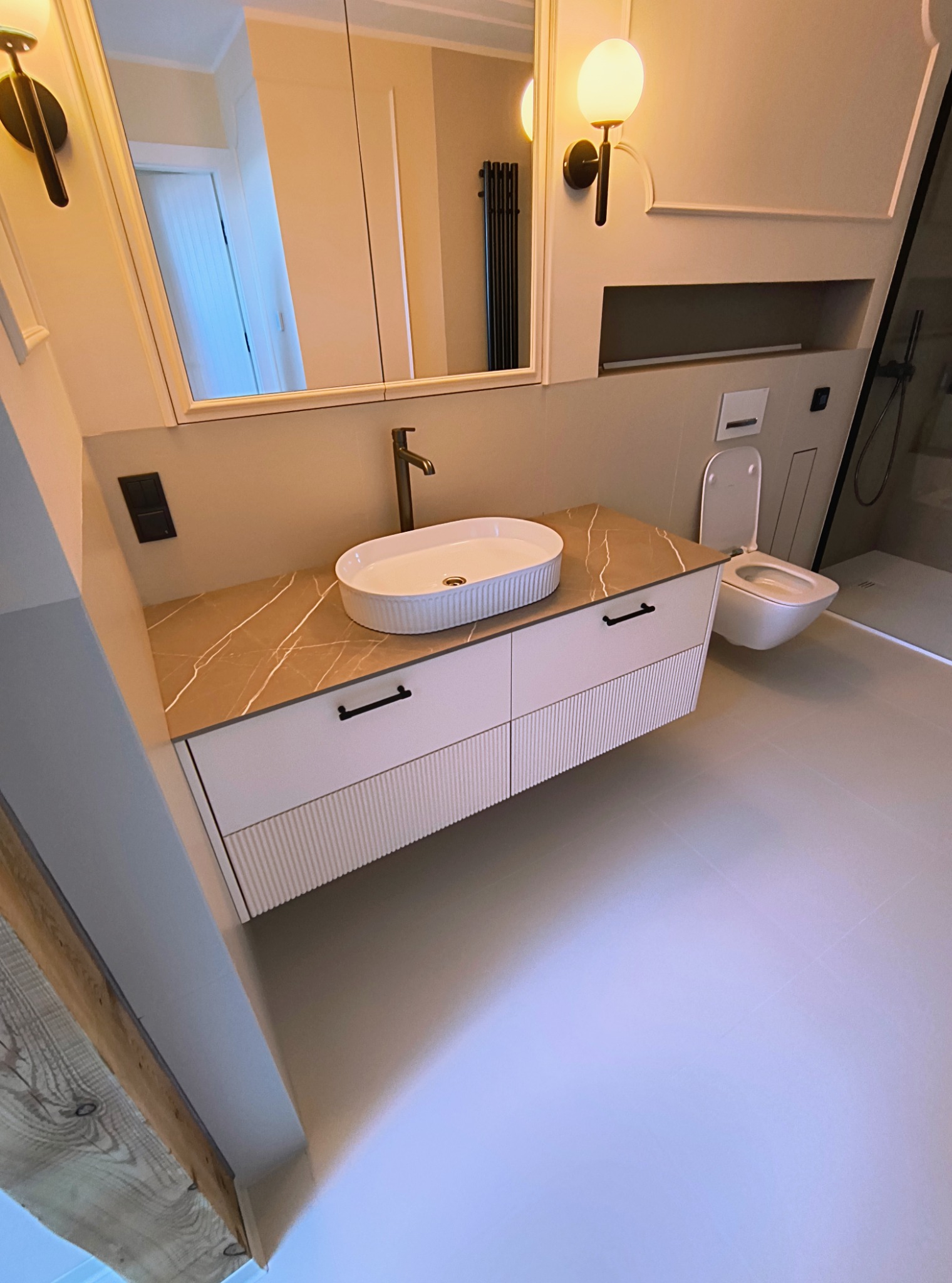 Nowoczesna łazienka z umywalką nablatową na szafce z ryflowanymi frontami, armatura w kolorze starego złota i beżowe płytki. Widoczna toaleta i fragment kabiny prysznicowej.