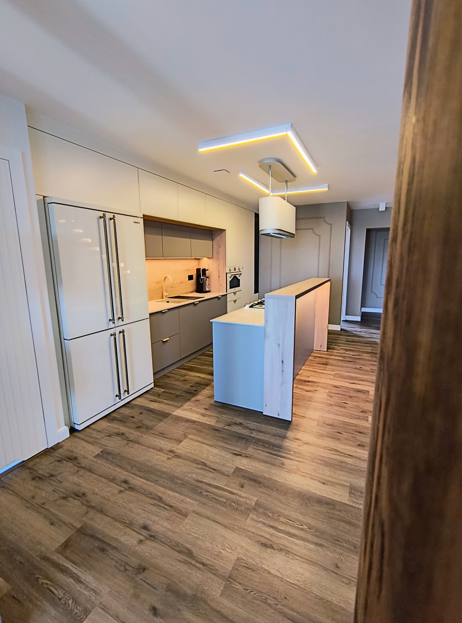 Nowoczesna kuchnia z wyspą, fronty w odcieniach szarości, drewniany blat i podłoga, oświetlenie LED. Widok z perspektywy wejścia, fragment zasłony na pierwszym planie.