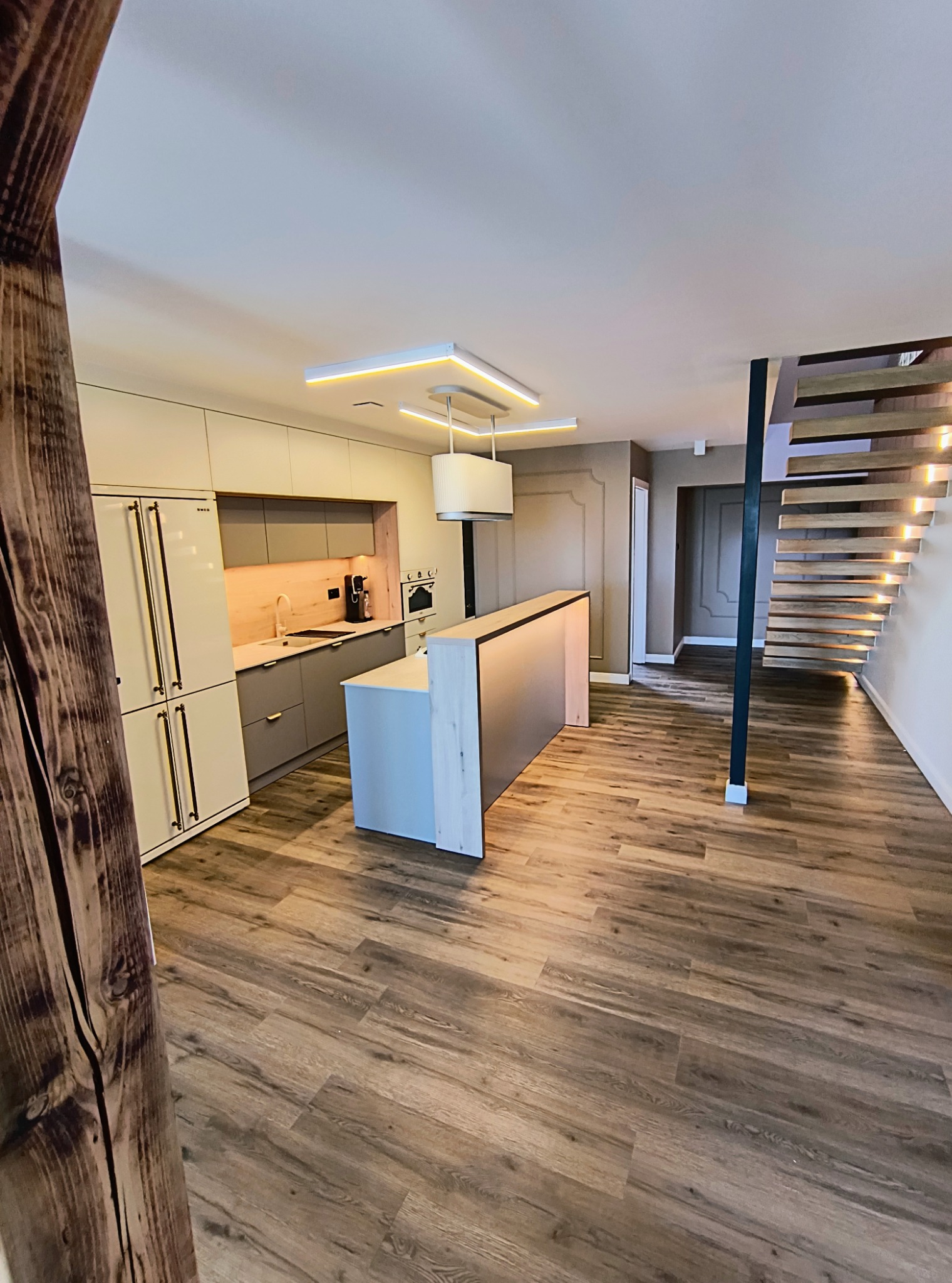 Nowoczesne wnętrze: kuchnia z wyspą i drewniane schody z podświetleniem, widok z perspektywy drewnianego słupa. Jasne kolory, minimalistyczny design.