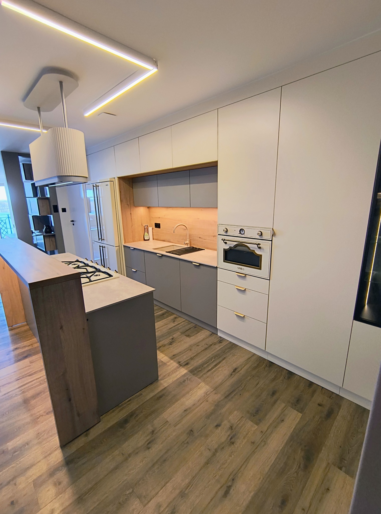 Nowoczesna kuchnia z wyspą, fronty w odcieniach szarości i bieli, drewniany blat wyspy, oświetlenie LED, podłoga imitująca drewno, piekarnik retro marki SMEG.