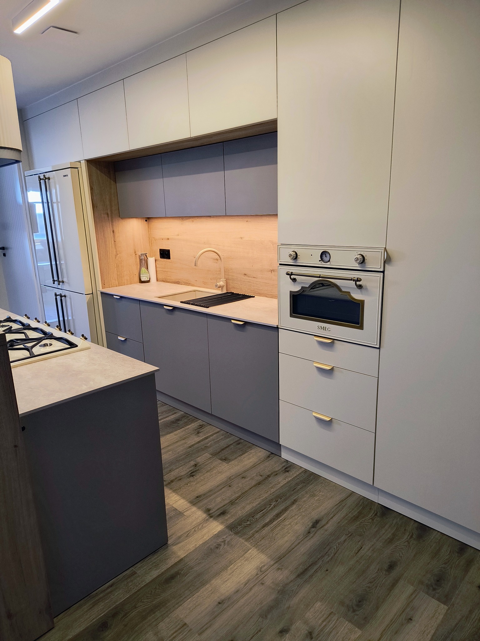 Nowoczesna kuchnia w odcieniach szarości i beżu, z drewnianym akcentem na ścianie i sprzętem AGD marki SMEG. Minimalistyczne fronty i drewniana podłoga.